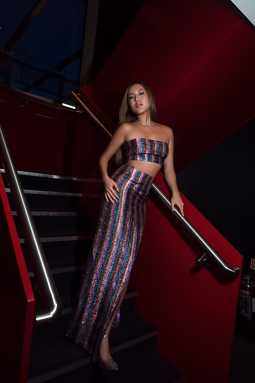 A woman posing on a red staircase wearing a matching two-piece set with vertical metallic rainbow stripes.