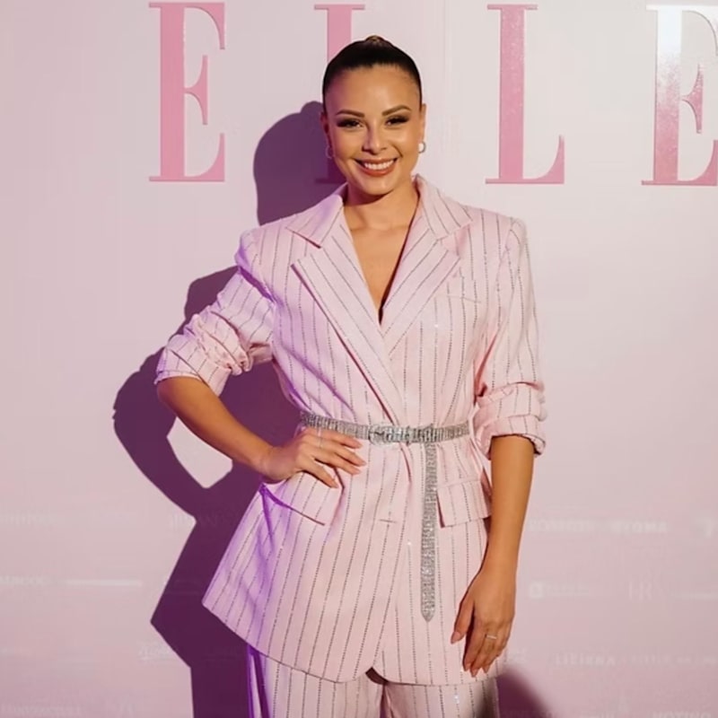 A woman posing in a light pink pinstripe blazer and matching trousers with a sparkly silver belt, standing against a pink background with 'ELLE' lettering.